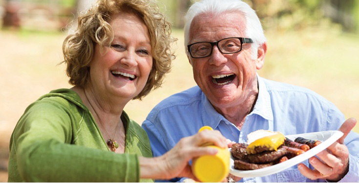 senior man and senior woman sitting at picnic table eating burgers and hot dogs