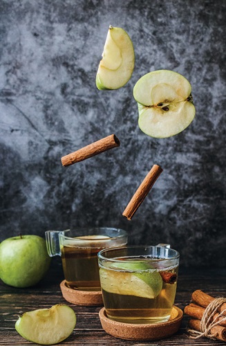 apple cider in short clear glass on table dressed with sliced apples and cinnamon sticks