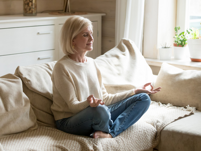 older woman in cream sweater, meditating on couch